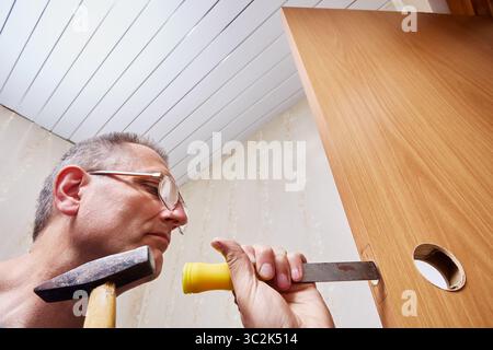 Intagliare una mortasa in una porta in legno, il personale di riparazione utilizza scalpello e martello per modellare con cura il foro per l'installazione della serratura durante il processo di montaggio della bulloneria della porta. Foto Stock