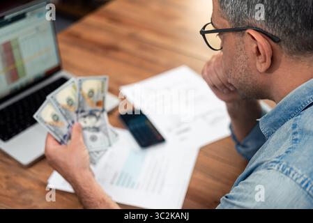 Dubbioso uomo dai capelli grigi guarda le banconote in dollari rispetto alle banconote non pagate impilate negli uffici commerciali Foto Stock