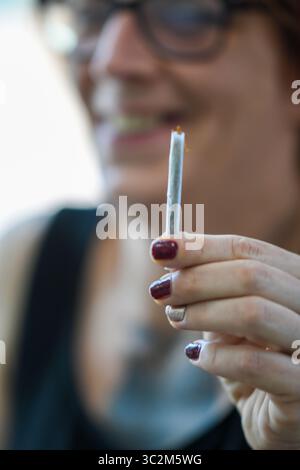 Cremona, Italia - 21 luglio 2025 Una donna sorridente con gli occhiali tiene una marijuana accesa tra le dita pronta a fumare durante la giornata all'aperto Foto Stock