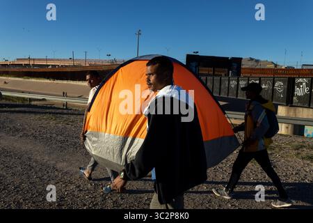 I migranti trasportano una tenda vicino al muro di frontiera di Ciudad Juarez sotto cieli limpidi, evidenziando la lotta e la resilienza in corso in mezzo a una complessa crisi migratoria. Foto Stock