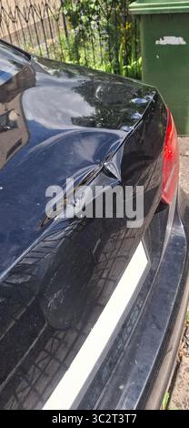 Foto ravvicinata del bagagliaio di un'auto nera che mostra segni visibili di ammaccature e graffi che necessitano di riparazione, con un cestino verde visibile sul marciapiede all'esterno Foto Stock