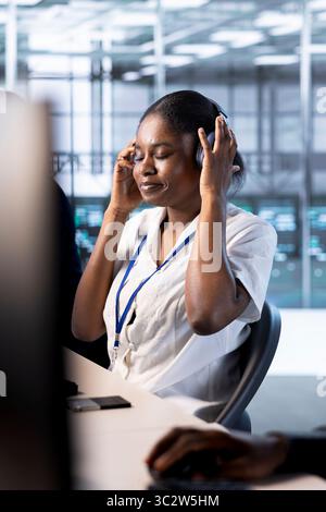 Amministratore afroamericano nel data center che indossa le cuffie e ascolta la musica durante l'esecuzione di test diagnostici di sistema. Donna in un server hub che ascolta canzoni e cerca problemi di rete che interessano i sistemi RIG Foto Stock