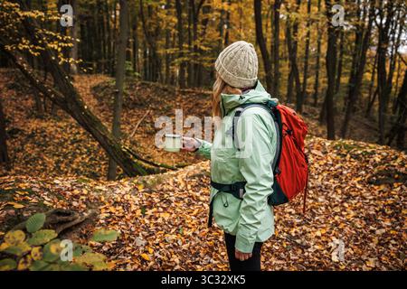 Donna in giacca impermeabile con zaino rosso che beve una bevanda calda nella tranquilla foresta autunnale. Escursioni nel bosco Foto Stock