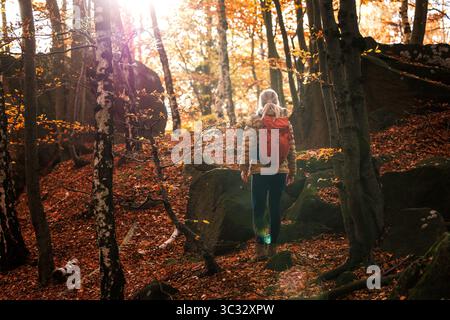 Donna che fa trekking nel bosco autunnale. Una turista con lo zaino rosso si sta arrampicando in salita. Vita attiva all'aperto Foto Stock