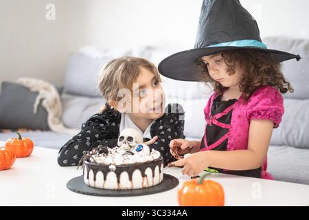 Two Girls in Halloween Costumes Decorating Spooky Cake Together Foto Stock