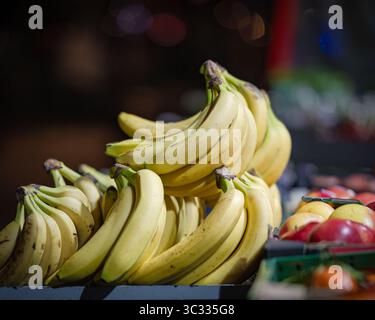 Primo piano di banane gialle mature in una bancarella di mercato di notte, con mele sfocate e altri frutti sullo sfondo. Foto Stock