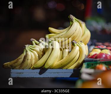 Primo piano di un mucchio di banane mature esposte su una banchina di legno in un mercato notturno. Mele e altri frutti sono parzialmente visibili sulla destra. Foto Stock