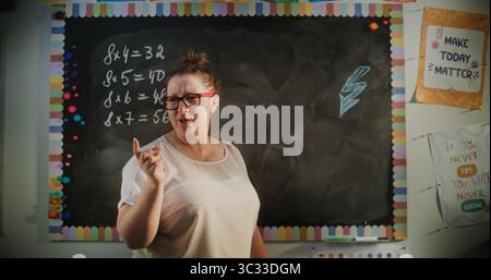 Vista posteriore della tabella delle moltiplicazioni per insegnanti femminili sulla lavagna, che istruisce gli studenti delle scuole primarie durante la lezione di matematica. Corso di matematica avanzata in classe moderna e colorata. Colpo statico. Foto Stock