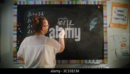 Vista posteriore dell'insegnante femminile che scrive sulla lavagna, spiegando la moltiplicazione e la divisione ai bambini delle scuole elementari durante la lezione di matematica. Corso di matematica in classe moderna. Colpo statico. Foto Stock