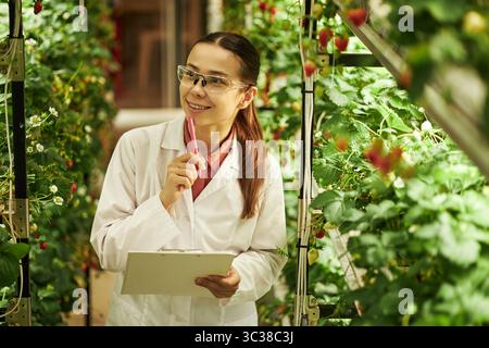 Giovane donna sorridente che indossa occhiali di sicurezza in piedi in serra tenendo appunti e penna, ispezionando le piante e prendendo appunti mentre è circondata da file di vegetazione coltivata Foto Stock
