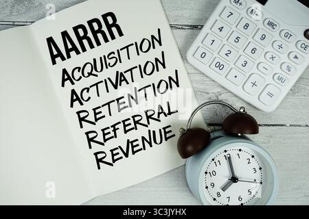 AARRR Acquisition, Activation, Retention, Referral, Revenue Text on paper notebook page with Alarm clock and calculator on Wooden background Foto Stock