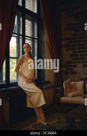 Una donna incinta siede comodamente a casa, cullando il suo bambino urtando contro una finestra. La luce solare calda filtra attraverso le tende, creando Foto Stock