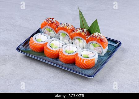 Sushi al salmone fresco con avocado, formaggio spalmabile e gamberi, disposti ad arte su un piatto blu con contorno e foglie verdi. Foto Stock