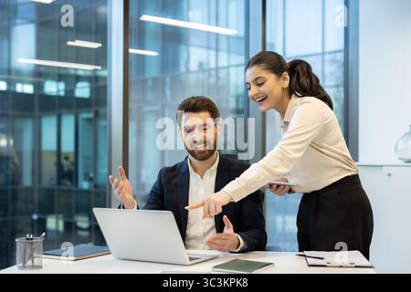 Una donna d'affari e un uomo d'affari stanno collaborando su un notebook in un ufficio moderno, condividendo un momento di discussione e un sorriso. Foto Stock