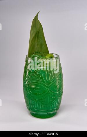 Un vivace cocktail verde servito in un bicchiere Tiki decorativo con una decorazione di foglie tropicali per un'esperienza di drink rinfrescante Foto Stock