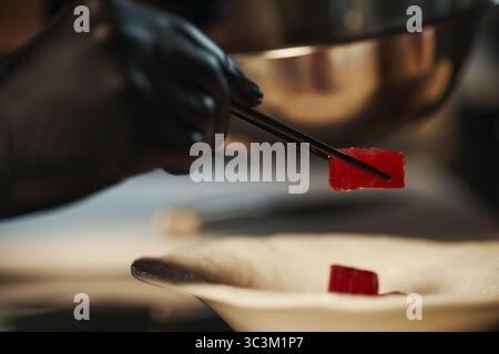 Primo piano di un adulto nero che indossa un guanto nero con bacchette per raccogliere un pezzo di tonno crudo sul piatto nella cucina del ristorante, preparando sushi o sashimi Foto Stock