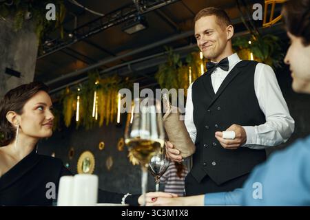 Giovane cameriere caucasico che serve vino alla giovane donna caucasica adulta e al giovane uomo caucasico adulto nel ristorante, tenendo in mano bottiglia di vino e cavatappi, sorridendo i clienti Foto Stock