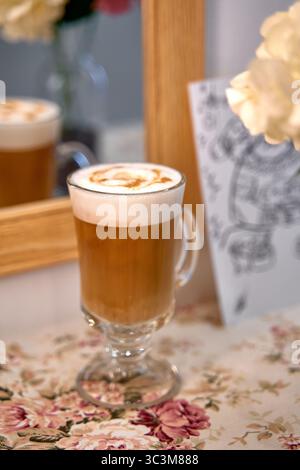 Un caldo bicchiere di delizioso caffè ricoperto da crema spuma e un tocco di caramello, splendidamente presentato su uno sfondo floreale e riflesso in una m Foto Stock