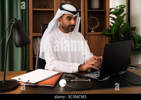 Uomo d'affari del Medio Oriente che indossa la kandura degli emirati che lavora in ufficio - uomo d'affari del Medio Oriente che lavora in kandura su un notebook alla volta Foto Stock