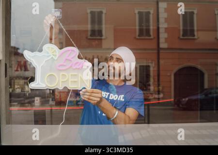 Cremona, Italia - 25 luglio 2025 giovane Sikh che appende un vibrante cartello al neon che dice aperto nella vetrina di un affascinante negozio che riflette la strada e. Foto Stock