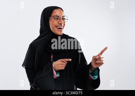 Ritratto in studio di una giovane donna musulmana allegra che indossa occhiali da vista e tradizionale abaya nera. Sorridendo e puntando le dita da parte durante lo showcasi Foto Stock