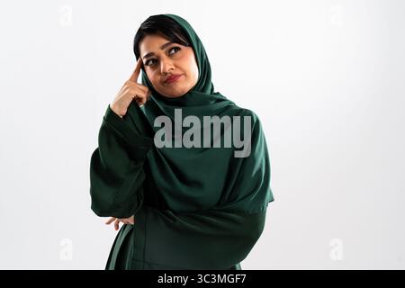 Ritratto in studio di una premurosa donna d'affari araba che indossa un hijab verde, toccando la testa con il dito mentre contempla una decisione contro una W. Foto Stock