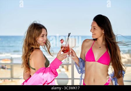 Due giovani donne, una con lunghi capelli castani e l'altra con capelli biondi ondulati, preparano cocktail colorati sorridendo gioiosamente al bar duri sulla spiaggia Foto Stock