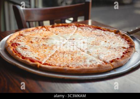 Una pizza al formaggio caldo appena sfornata si siede su un tavolo in un accogliente ristorante di Brooklyn, USA. Il delizioso aroma del formaggio fuso riempie l'aria, Foto Stock