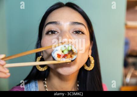 Una donna tiene un pezzo di sushi con bacchette, assaporando l'esperienza in un vivace ristorante di sushi. La sua espressione cattura la gioia di cenare fuori Foto Stock