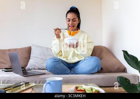 Una donna latina si rilassa a casa, sedendosi comodamente sul divano con una ciotola in mano. Intorno a lei ci sono oggetti di uso quotidiano come un notebook e una tazza, illustratore Foto Stock