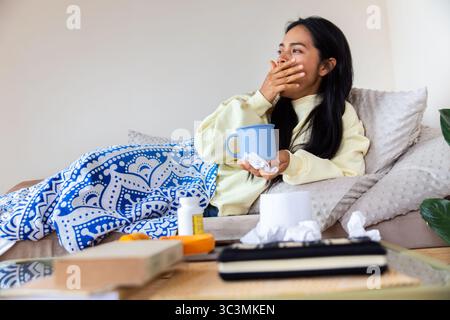 Una donna latina che si sente male, seduta su un divano a casa. Avvolta in una coperta, tiene una bevanda calda. Intorno a lei ci sono tessuti e medicine, conve Foto Stock