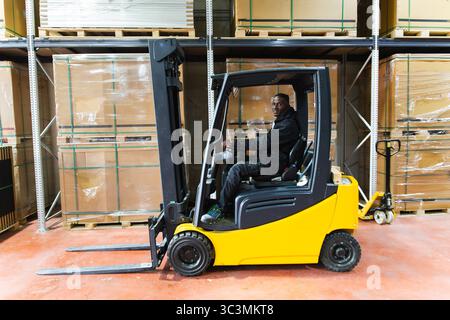 Un uomo gestisce un carrello elevatore giallo in un magazzino di pannelli solari, circondato da pallet e scatole di cartone. È seduto e concentrato sulla manovra dell'eq Foto Stock