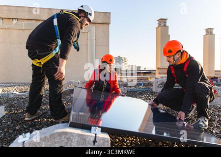 Tecnici esperti che indossano attrezzature di sicurezza lavorano insieme per installare pannelli solari su un tetto, mostrando soluzioni di energia rinnovabile in un ambiente urbano Foto Stock