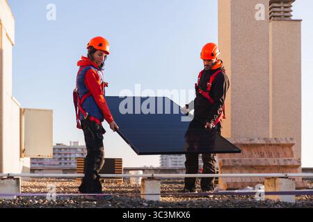 I lavoratori che utilizzano attrezzature di sicurezza installano pannelli solari sul tetto, garantendo un cablaggio energetico efficiente. Il tramonto offre uno sfondo sereno, evidenziando su Foto Stock
