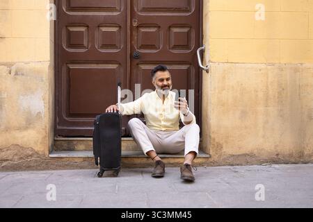 Un uomo indiano siede su un gradino di pietra, controllando il suo smartphone mentre si riposa accanto alla sua valigia. È vestito con un abbigliamento estivo leggero, godendosi un soleggiato Foto Stock