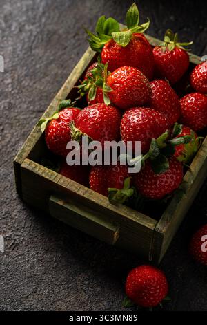 Una scatola di legno rustica piena di fragole rosse vivaci collocate su una superficie scura e ruvida. Le fragole, fresche e succose, mostrano il loro lucido sk Foto Stock