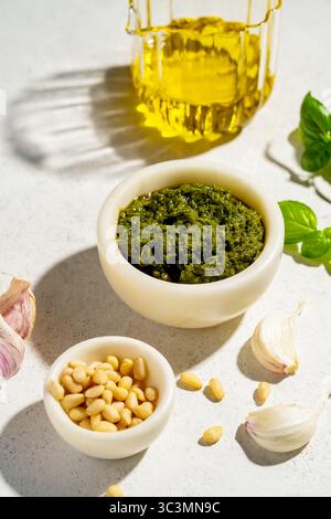 Dall'alto, l'immagine cattura il pesto in una ciotola bianca, pinoli e aglio su una superficie bianca. Un contenitore in vetro di olio d'oliva e foglie di basilico sono disponibili Foto Stock