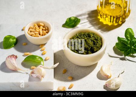 Dall'alto, uno sfondo in marmo mostra una ciotola di pesto verde, pinoli, spicchi d'aglio, foglie di basilico e un vaso di olio d'oliva, che incarna il Mediterraneo Foto Stock