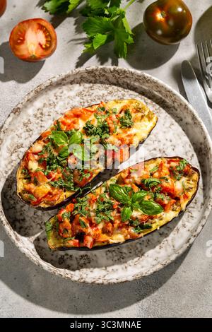 Metà di melanzane deliziosamente sfornate ripiene di verdure fresche ed erbe aromatiche, guarnite con basilico, esposte su un piatto rustico macchiato, pronte Foto Stock
