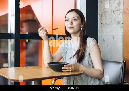 Una donna siede in un elegante caffè, assaporando una deliziosa cheesecake a colazione. Tiene in mano un cucchiaio e si siede a un tavolo di legno, gustando il suo pasto Foto Stock