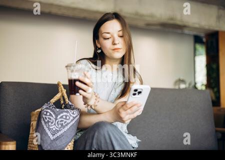 Una donna siede comodamente in un accogliente bar, tenendo un caffè in una mano e il suo smartphone nell'altra. Sembra rilassata, coinvolgente con il suo dispositivo. Foto Stock