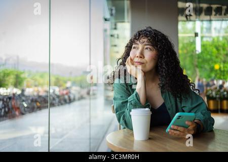 Una giovane donna con i capelli ricci siede in un bar moderno, tenendo in mano uno smartphone e guardando fuori. Una tazza di caffè poggia sul tavolo, circondato da un tranquillo urba Foto Stock