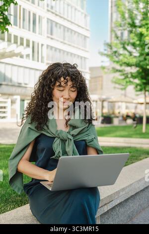 Una giovane donna con i capelli ricci lavora sul suo computer portatile mentre è seduta su una panchina in un parco urbano. È concentrata e gode dei tranquilli dintorni all'aperto Foto Stock