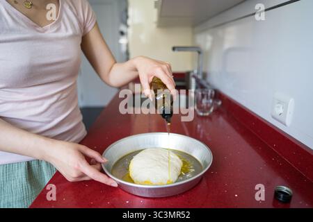 Donna della cucina moderna che versa olio d'oliva sull'impasto della focaccia in una teglia da forno. Si concentra sulla preparazione del tradizionale pane italiano con cura e Foto Stock