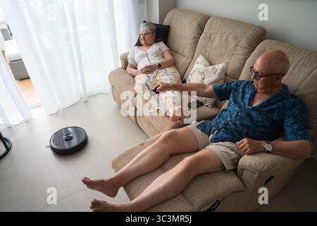 Una coppia anziana si siede comodamente sul divano, godendosi la facilità di un aspirapolvere robot intelligente nella loro casa moderna. Adottare la tecnologia per un AN attivo Foto Stock