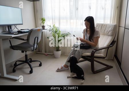 Una donna asiatica siede in un ufficio domestico con il suo cane, mentre tiene un aspirapolvere robot in grembo. La camera è luminosa e moderna, con una scrivania per computer e PL Foto Stock