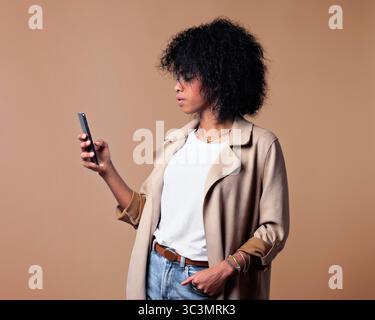 Donna nera in un outfit casual incentrato sullo smartphone, indossa un cappotto beige, top bianco e jeans, con un'espressione neutra su uno sfondo semplice. Foto Stock