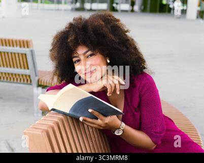 Una donna egiziana sorridente ama leggere un libro seduto comodamente su una panchina all'aperto. La sua posa rilassata e l'ambiente piacevole suggeriscono un amore Foto Stock