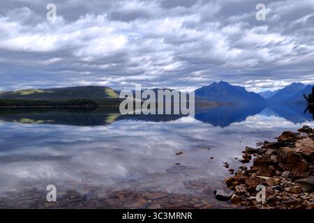 La costa rocciosa incontra le acque limpide del lago McDonald che riflettono picchi carichi di nuvole e colline boscose illuminate dal sole nel tranquillo rifugio del Glacier National Park. Foto Stock