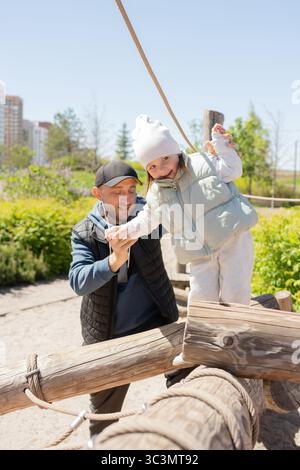 Un padre sostiene sua figlia mentre si bilancia su una struttura di legno in un parco. Si godono una giornata di sole all'aperto, sorridono e spendono qualità Foto Stock